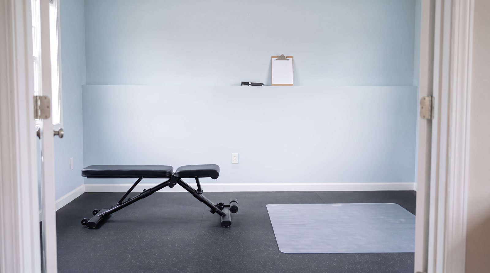 A view of an empty workout bench and separate mat area through a doorway.