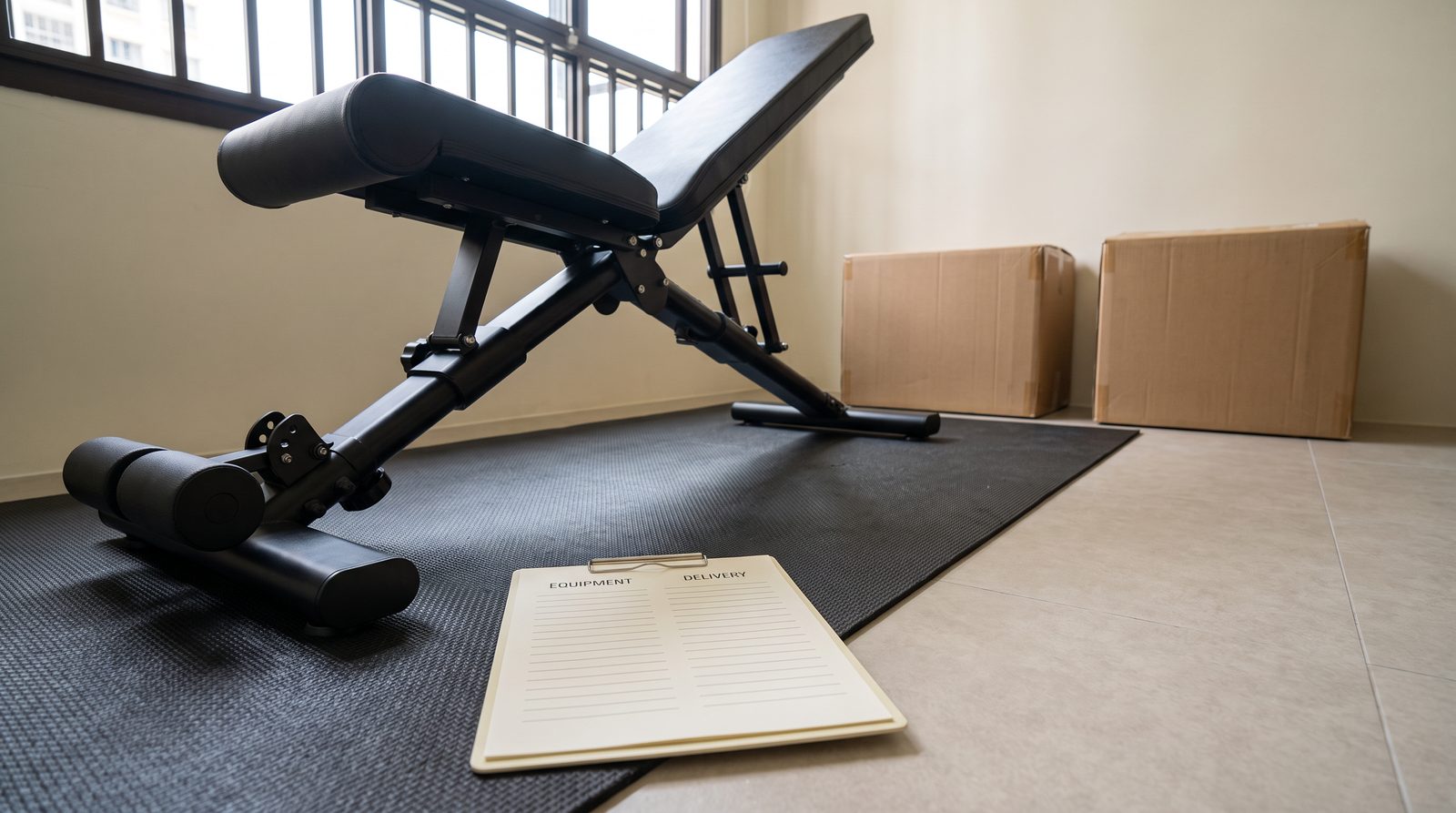 An inclined exercise bench on floor protection beside cartons and a blank equipment and delivery checklist.